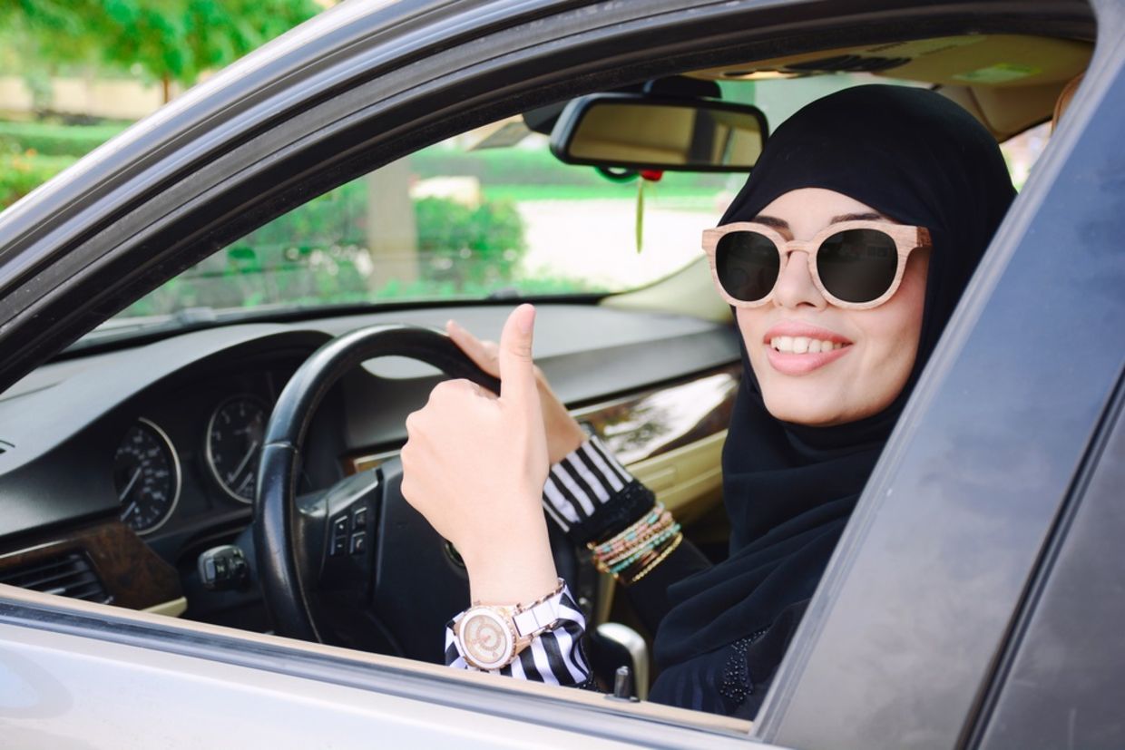 Happy business arabic lady in dark abaya and hijab driving her car and showing thumb up. Smiling arabian lady in sunglasses taking steering wheel of her new car.