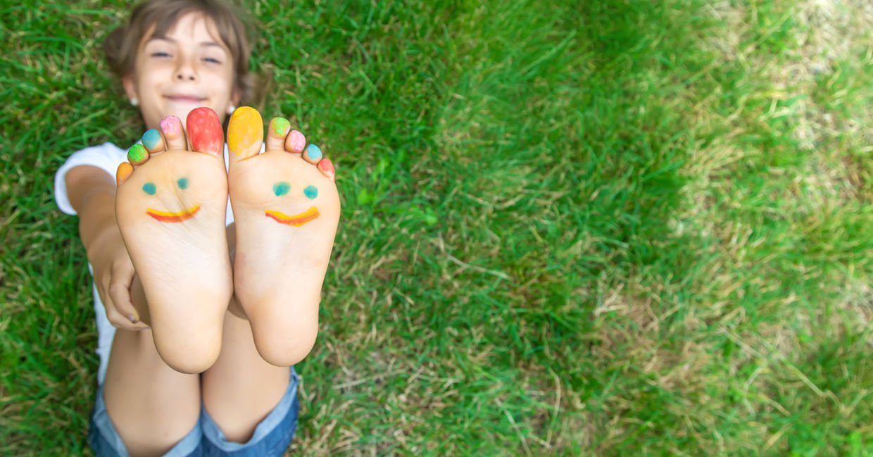 A child lies on the grass, feet up, with painted toes and a happy face on her feet.
