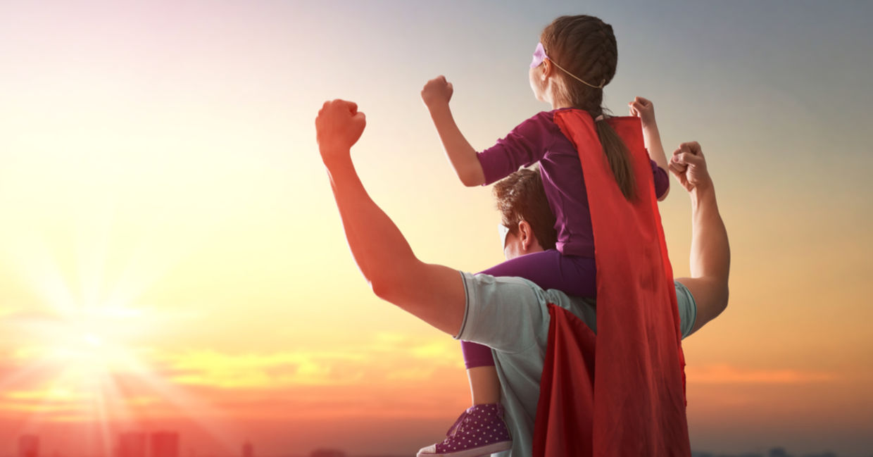 A father holds his daughter on his shoulders, both of them wearing empowering superhero capes.