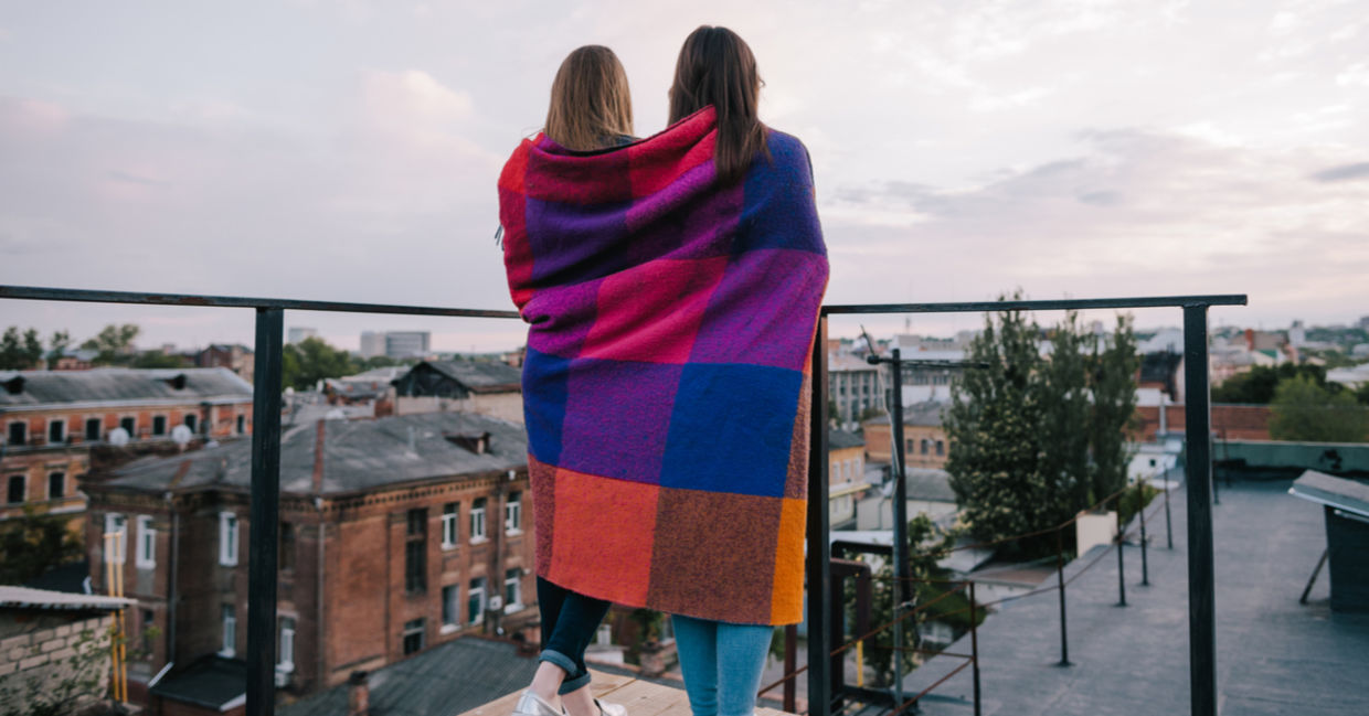 Two friends wrapped in a blanket stand outside on a balcony.