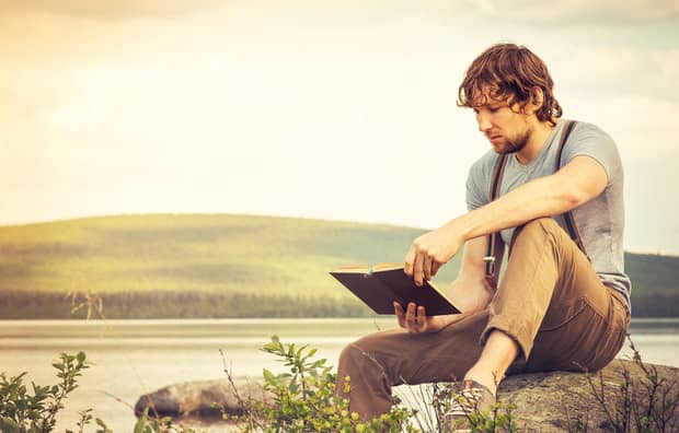 A young man with curly hair and a beard sits on a rock reading a book, wearing a gray tank top and khaki pants with a backpack, overlooking a serene lake and rolling hills in warm golden light.
