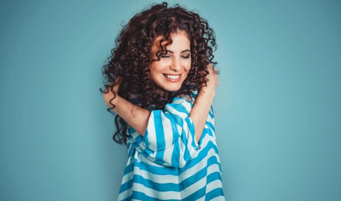 Woman with dark curly hair wearing a blue and white striped shirt, smiling with hands near her head against a turquoise background.