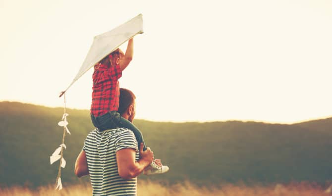 Child on parent's shoulders flying a white kite at sunset with rolling hills in the background during golden hour.