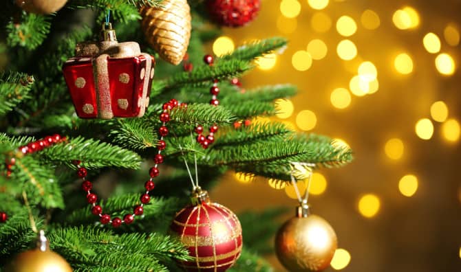 Decorated Christmas tree with red and gold ornaments, beaded garland, and gift box ornament with blurred golden lights in the background.