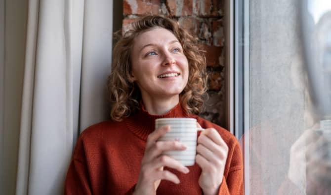 A smiling woman next to a window holds a mug.
