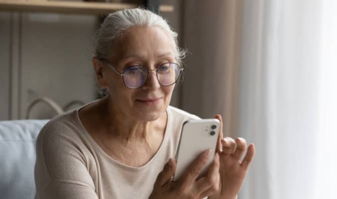An older woman in eyeglasses sits on a couch at home and holds a smart phone.