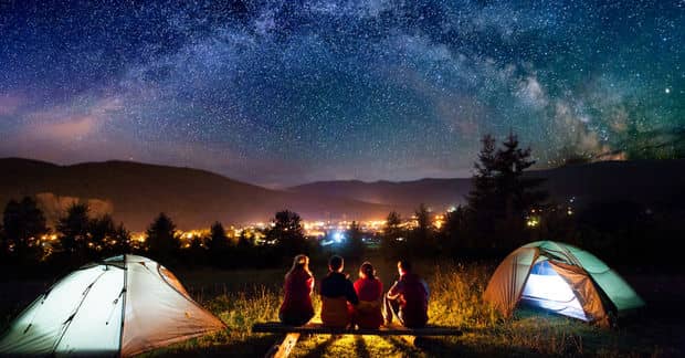 Group of people around a campfire between glowing tents at night, with the Milky Way and starry sky visible above silhouetted mountains.