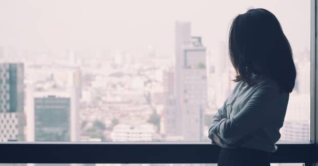 A woman stands silhouetted by a window, arms crossed, gazing out over a hazy urban cityscape of buildings.