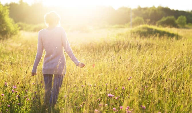 Person in a pink sweater walking through a wildflower field at sunset with golden hour lighting and distant trees.