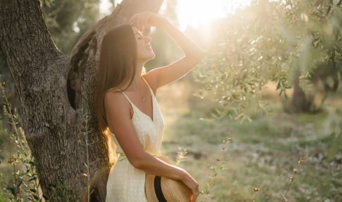 A woman in a yellow dress enjoys the warm sunlight while leaning against a tree.
