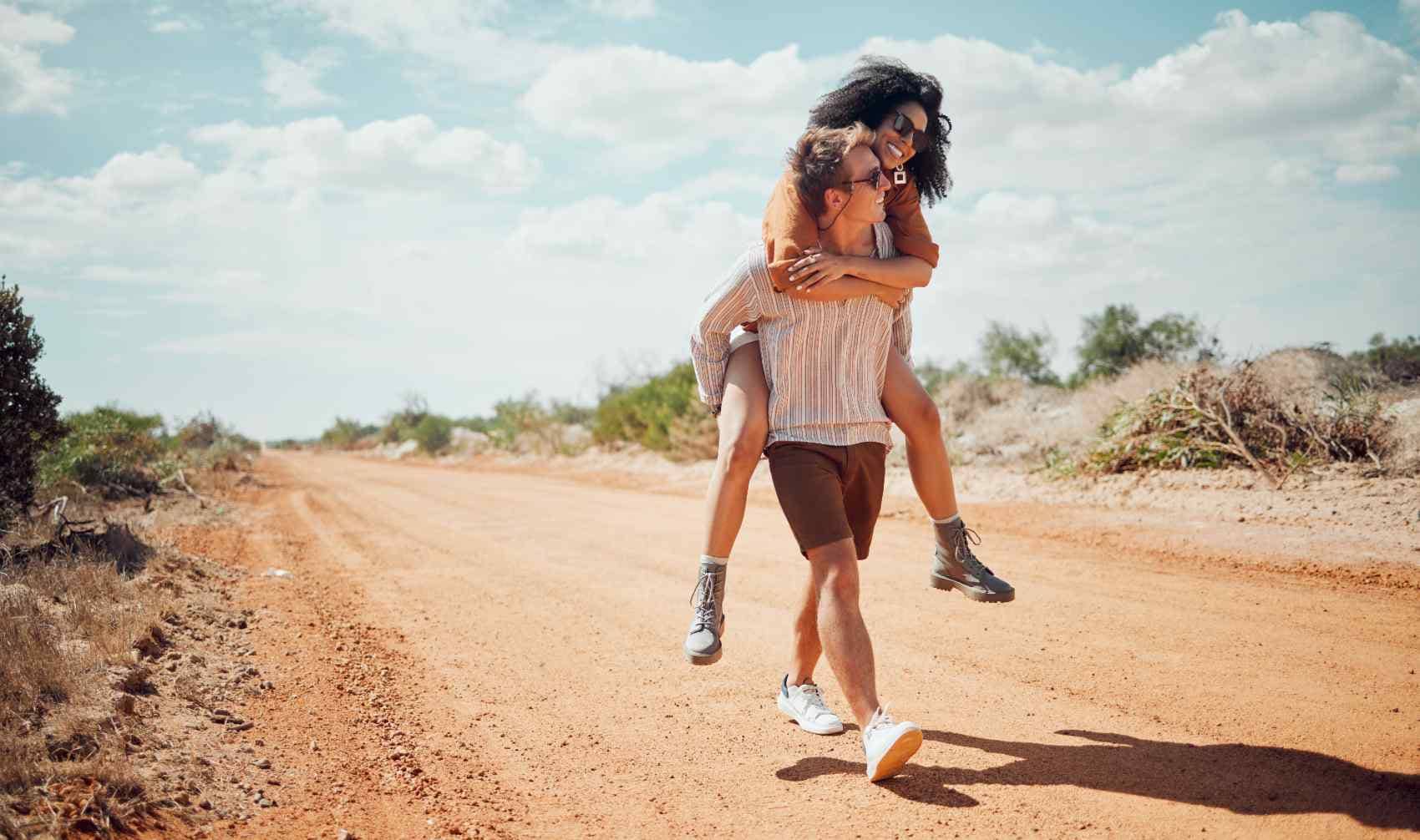 A young man gives a woman a piggyback ride on a desert road.