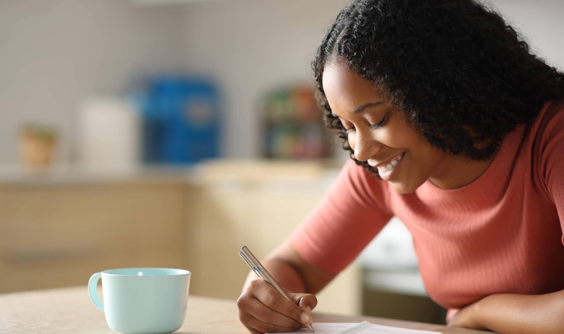 A happy woman writes in an agenda while sitting in the kitchen at home.