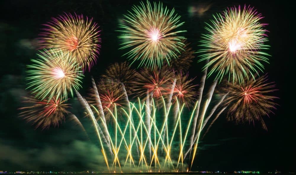 Majestic fireworks with green and pink bursts illuminate the night sky, creating a stunning spectacle over the water.
