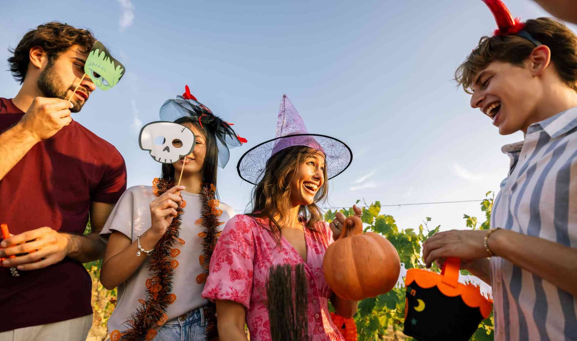 A smiling woman wearing a witch hat holds a pumpkin while her friends in Halloween costumes laugh and interact together outdoors in a vineyard.