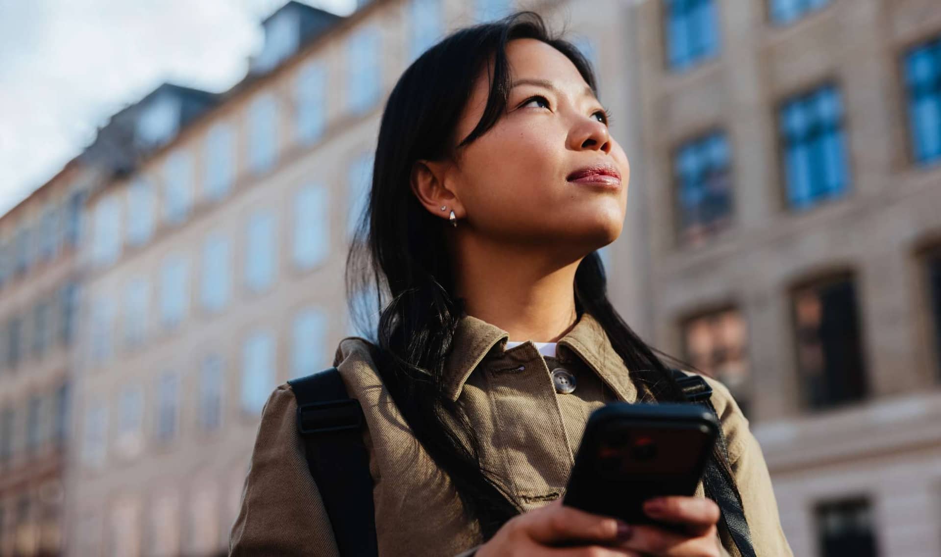 A young woman holds a phone and looks up on a city street.