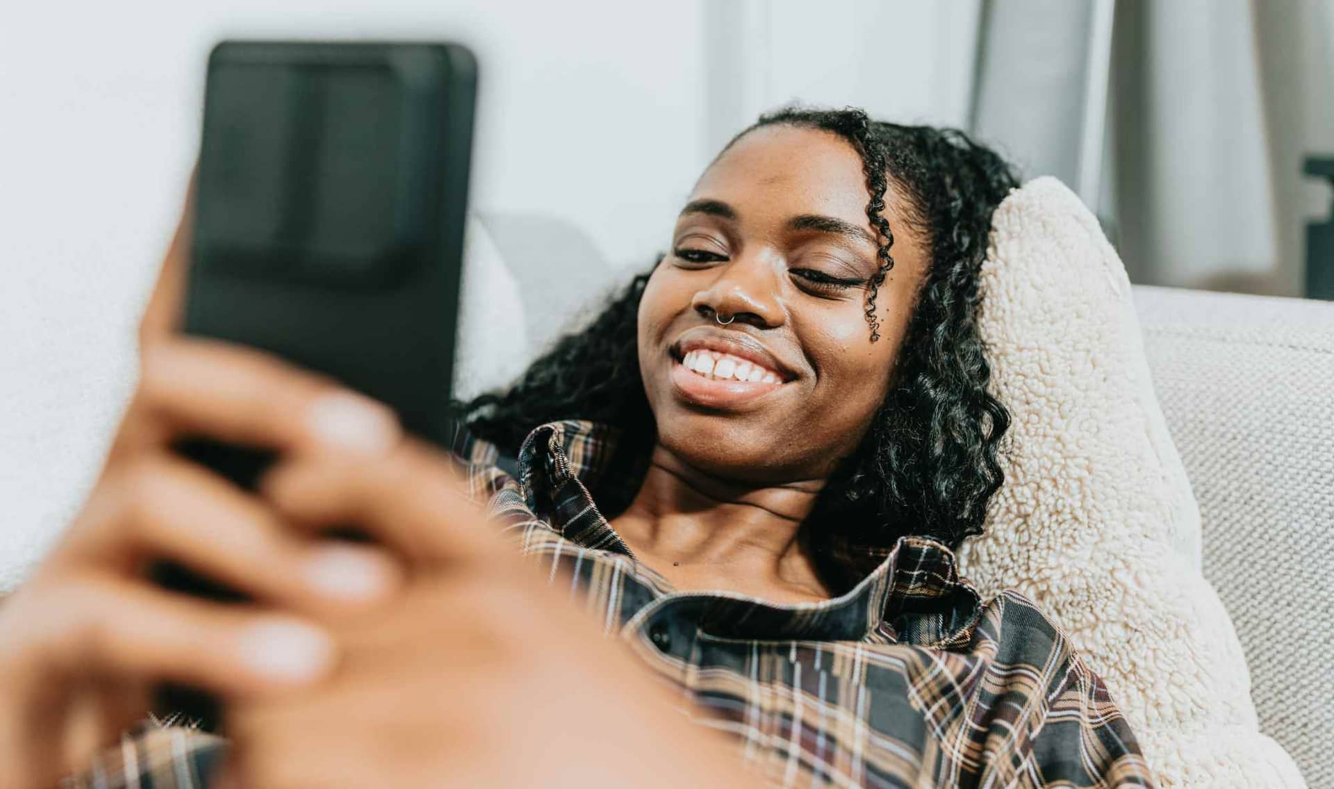 A smiling woman relaxing on a couch while looking at her smartphone.