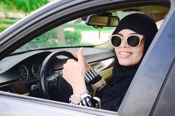 Happy business arabic lady in dark abaya and hijab driving her car and showing thumb up. Smiling arabian lady in sunglasses taking steering wheel of her new car.