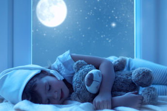 A little girl hugs a teddy bear with a full moon shining outside her window.
