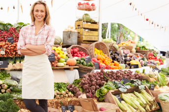 A vendor stands in front of her vegetable stand at an outdoor market.