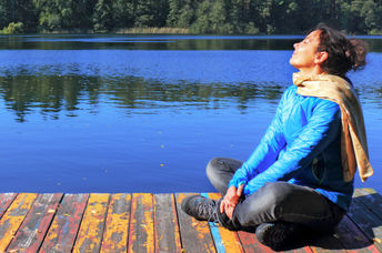 A woman sitting on a dock by a lake is doing a breathing exercise to help tone her vagus nerve.