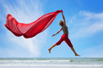 A happy, fulfilled woman jumps on the beach, trailing a bright red scarf high in the air.
