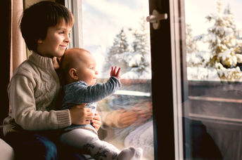 Boy holding his baby brother, sitting by the window in living room, looking at a snowy landscape outdoors.