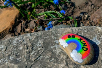 The words be kind and a rainbow are painted on a pebble with spring blue flowers in the background.