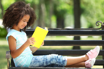 A young girl enjoys reading her book on a park bench.