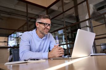 A man wearing glasses smiles while looking at a laptop screen.