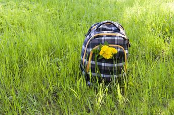 A school backpack decorated with dandelions stands among the fresh high grass during a hike.