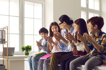 A female teacher claps together with a group of happy children.