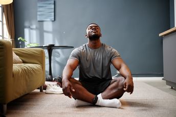 A young adult man sits cross-legged on a carpet in a living room.