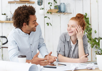 Beautiful young redhead European woman designer with hair bun laughing at jokes of her cheerful charismatic dark-skinned male colleague