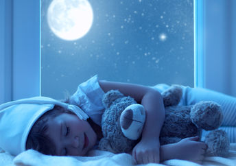 A little girl hugs a teddy bear with a full moon shining outside her window.
