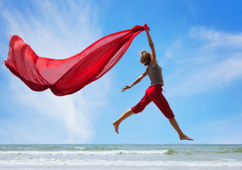 A happy, fulfilled woman jumps on the beach, trailing a bright red scarf high in the air.