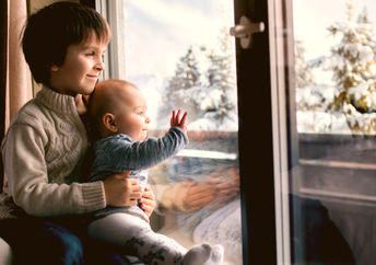 Boy holding his baby brother, sitting by the window in living room, looking at a snowy landscape outdoors.