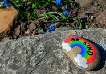The words be kind and a rainbow are painted on a pebble with spring blue flowers in the background.