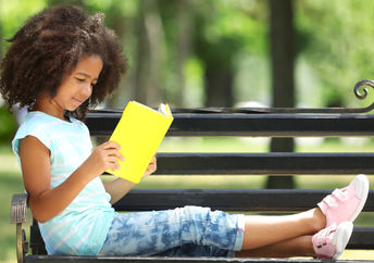 A young girl enjoys reading her book on a park bench.
