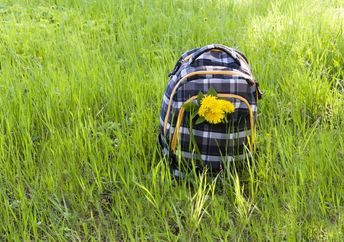 A school backpack decorated with dandelions stands among the fresh high grass during a hike.