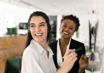 Two female coworkers smiling as they prepare to write on a whiteboard.