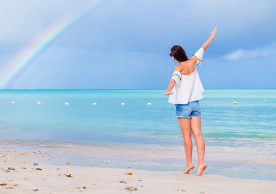 A young woman joyfully walks along a beach with a rainbow on the horizon.