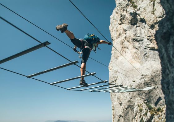 A person balances on a metal rope bridge against a blue sky and mountain.