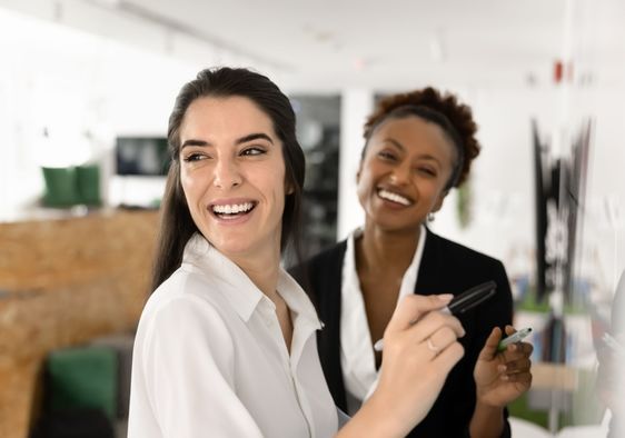 Two female coworkers smiling as they prepare to write on a whiteboard.
