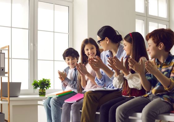 A female teacher claps together with a group of happy children.