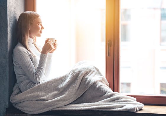 A woman sits by a window and holds a cup.
