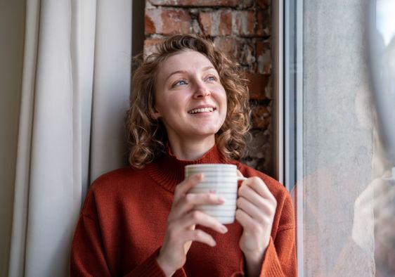 A smiling woman next to a window holds a mug.