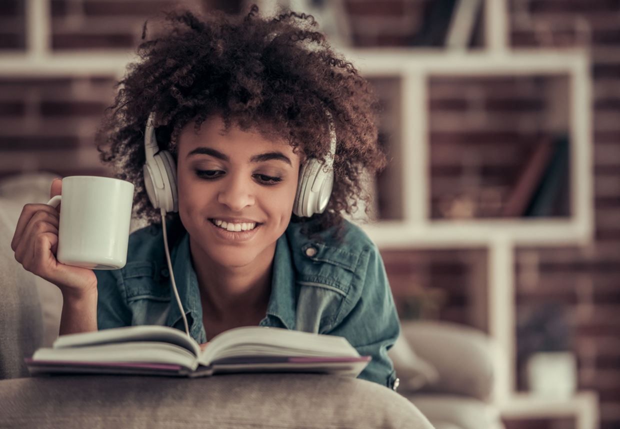Afro American girl in headphones is listening to music, reading a book, drinking coffee