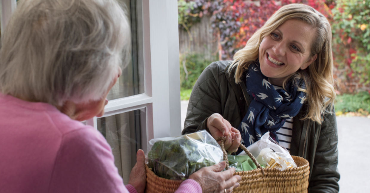 A smiling woman reaches out by delivering a basket of food to a senior.