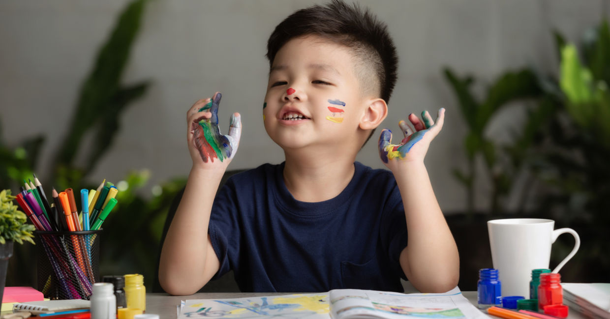 A young boy happily painting with paint all over his hands and face.