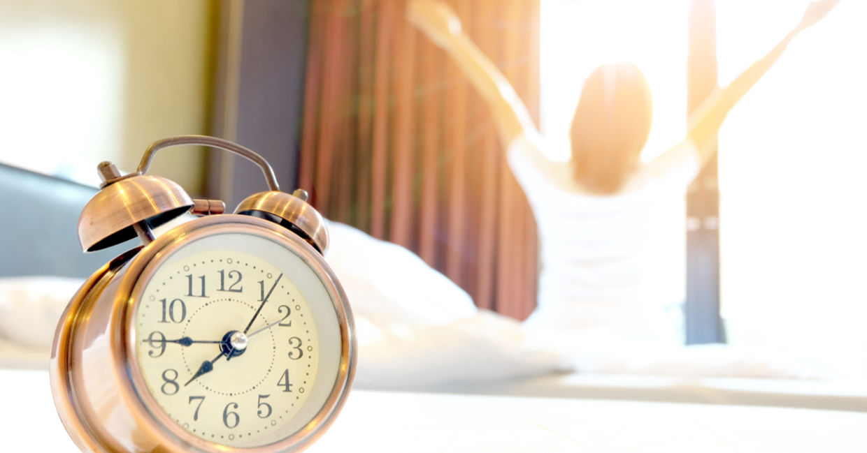 An alarm clock in the bedroom with a woman waking up early looking refreshed.